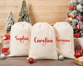 May include: Three off-white drawstring gift bags with red text that reads "Sophia", "Caroline", and "Susanne", each with a heart symbol. The bags are on a wooden surface, with Christmas decorations in the background.