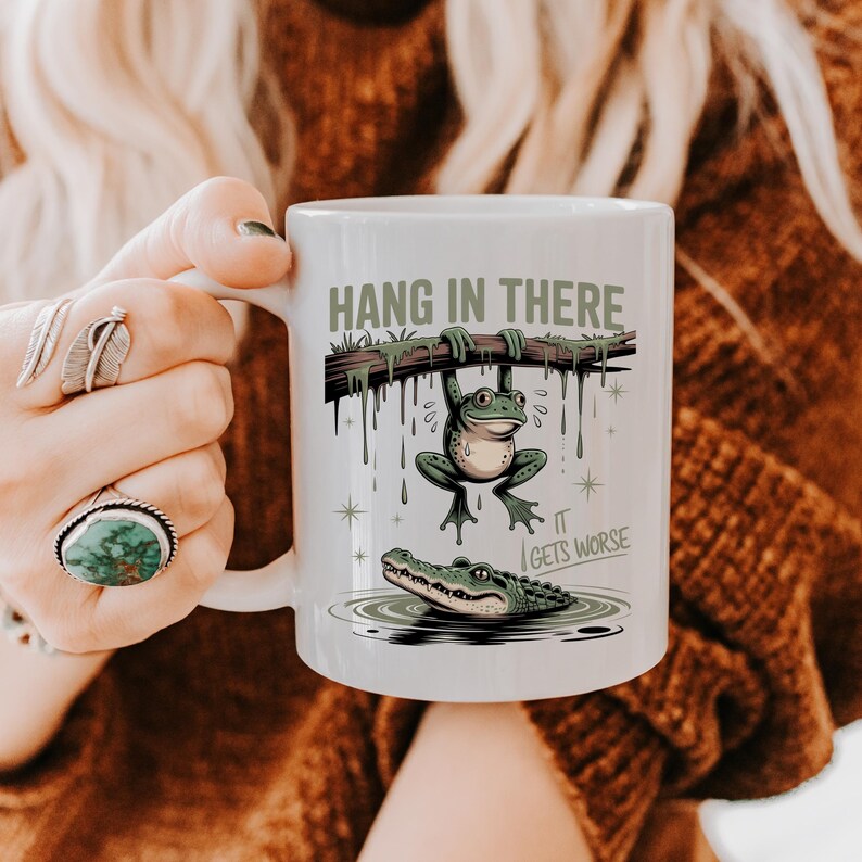 May include: White ceramic mug with a humorous illustration. The design features a frog hanging from a branch above an alligator in water, with the text "HANG IN THERE" and "IT GETS WORSE".