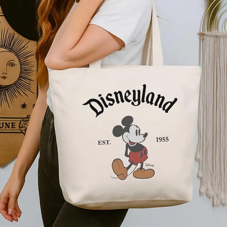 Op de afbeelding: Cr&egrave;mekleurige draagtas met het Disneyland-logo in zwart script, Mickey Mouse en de tekst "EST. 1955". De tas heeft een eenvoudig ontwerp met lange handvatten om over de schouder te dragen.