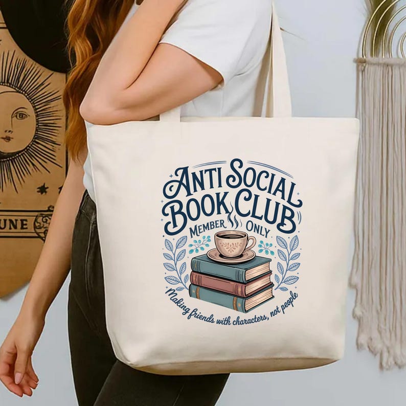 May include: Cream-colored tote bag featuring the text "Anti Social Book Club Member Only" in a whimsical font. The design includes a stack of books, a coffee cup, and the phrase "Making friends with characters, not people."