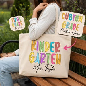 May include: A beige canvas tote bag featuring the word KINDERGARTEN in large, multi-colored brushstroke-style lettering with the name Mrs. Taylor printed in black script below. Two smaller square graphics show custom text options.