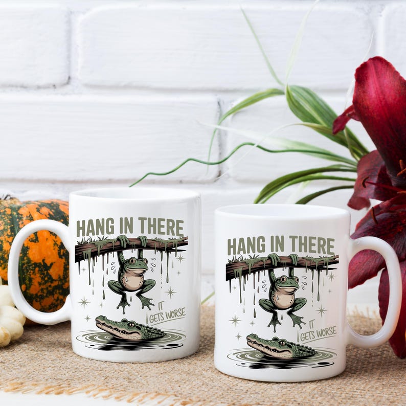 May include: Two white ceramic mugs with a humorous design. The mugs feature a cartoon frog hanging from a branch above an alligator in water. The text "HANG IN THERE" and "IT GETS WORSE" are printed on the mugs.