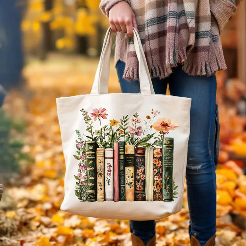 May include: Cream-colored tote bag featuring a floral and book design. The design includes colorful flowers and a row of vintage-style books. The bag has sturdy handles for carrying and is held by a person.