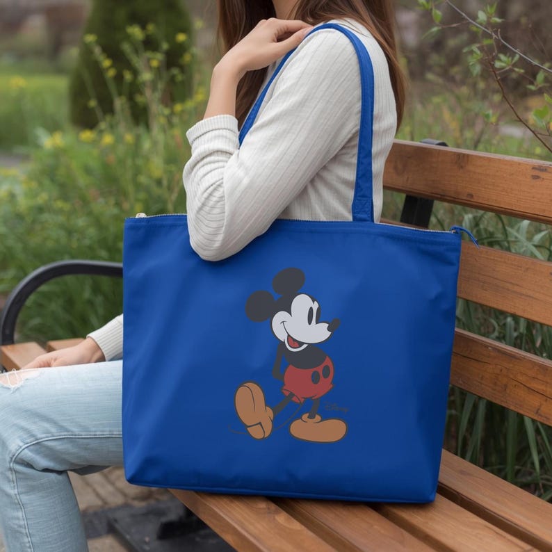 Puede incluir: Un bolso tote azul real con un gr&aacute;fico de Mickey Mouse. El bolso tiene cierre de cremallera y correas azules. El personaje de dibujos animados est&aacute; representado con sus ic&oacute;nicos pantalones cortos rojos, zapatos amarillos y orejas negras.