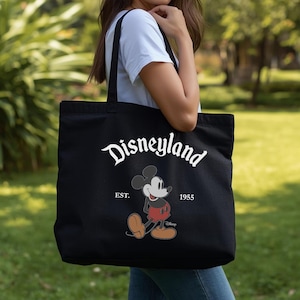 Op de afbeelding: Zwarte draagtas met witte "Disneyland"-tekst en een Mickey Mouse-afbeelding. De tas bevat ook de tekst "EST. 1955". De tas wordt vastgehouden door een persoon in een wit t-shirt en een blauwe spijkerbroek.