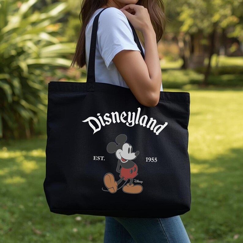 Op de afbeelding: Zwarte draagtas met witte "Disneyland"-tekst en een Mickey Mouse-afbeelding. De tas bevat ook de tekst "EST. 1955". De tas wordt vastgehouden door een persoon in een wit t-shirt en een blauwe spijkerbroek.