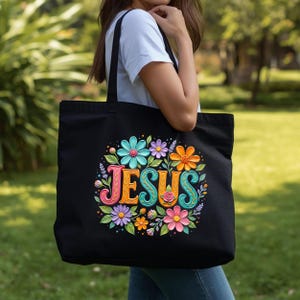 May include: Black tote bag featuring the word "JESUS" in colorful, stylized letters surrounded by a floral design. The flowers are in shades of blue, orange, pink, and purple. The bag is carried by a person.