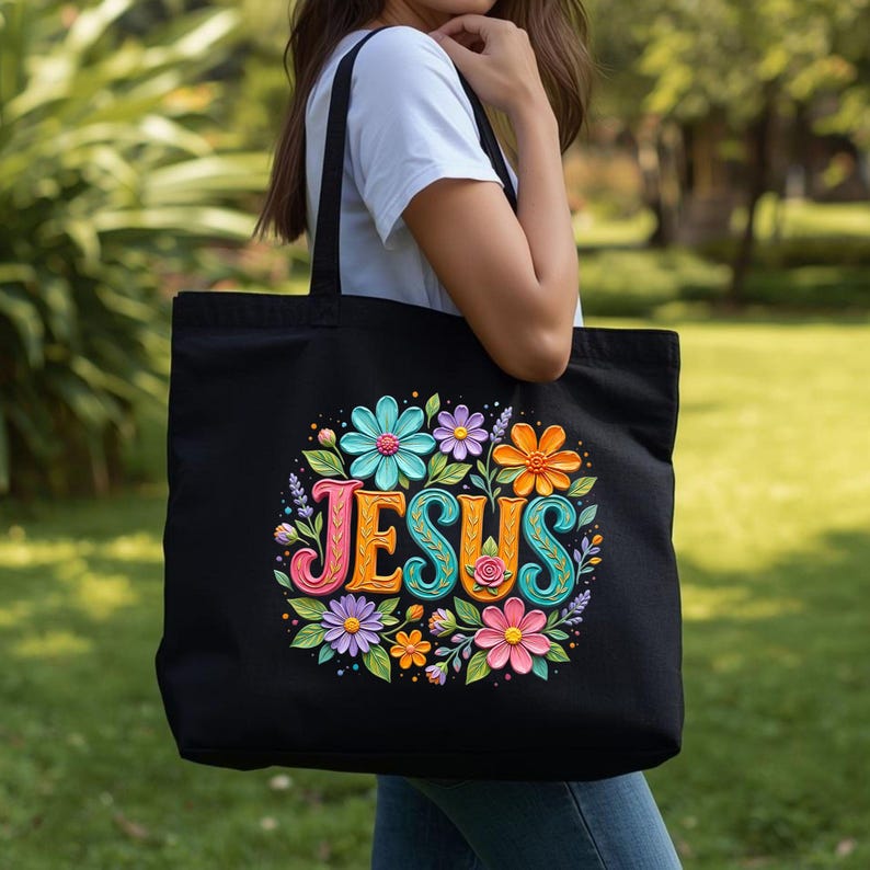 May include: Black tote bag featuring the word "JESUS" in colorful, stylized letters surrounded by a floral design. The flowers are in shades of blue, orange, pink, and purple. The bag is carried by a person.