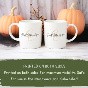 May include: Two white ceramic mugs with the text "Print Studio Art" in black and gold script. The mugs have a curved handle and are displayed in front of two orange pumpkins. The text "PRINTED ON BOTH SIDES" is visible.