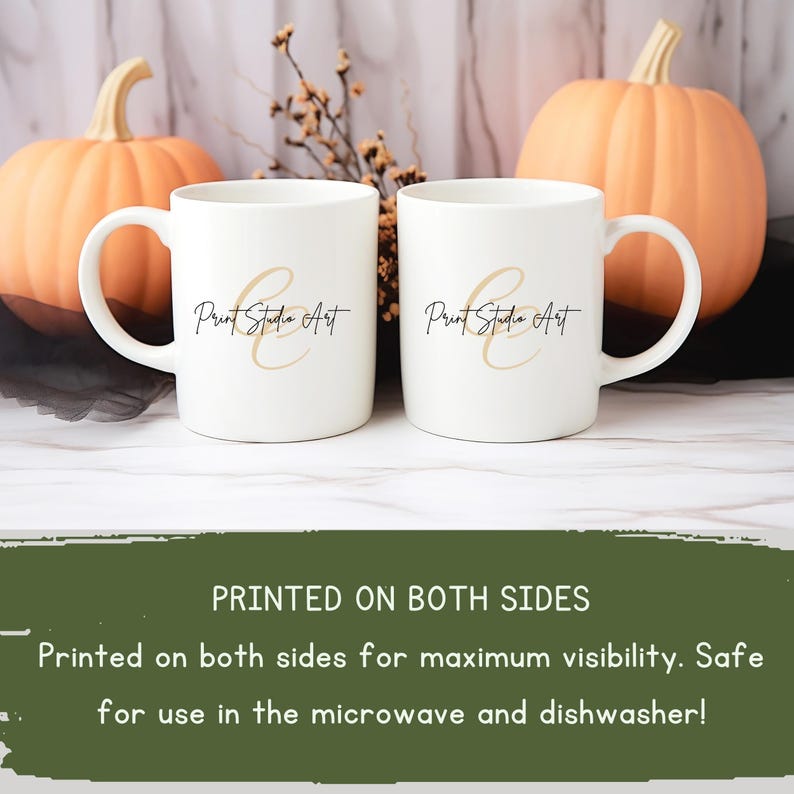 May include: Two white ceramic mugs with the text "Print Studio Art" in black and gold script. The mugs have a curved handle and are displayed in front of two orange pumpkins. The text "PRINTED ON BOTH SIDES" is visible.