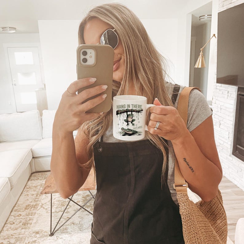 May include: White ceramic mug with the text "HANG IN THERE" and a cartoon frog design. The mug features a frog hanging from a branch, with a second frog on a lily pad. The mug has a white handle.