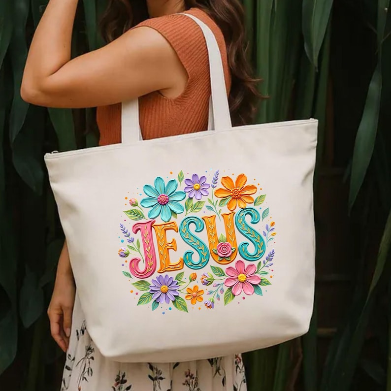May include: Cream-colored tote bag with the word "JESUS" in colorful, stylized letters surrounded by a floral wreath. The flowers are in shades of blue, orange, purple, and pink, with green leaves. The bag has white handles and a zipper closure.