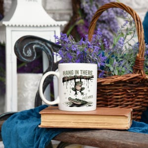 May include: White ceramic mug with the text "HANG IN THERE" and a cartoon frog hanging from a branch above an alligator. The phrase "IT GETS WORSE" is below the alligator. The mug is on a stack of books.