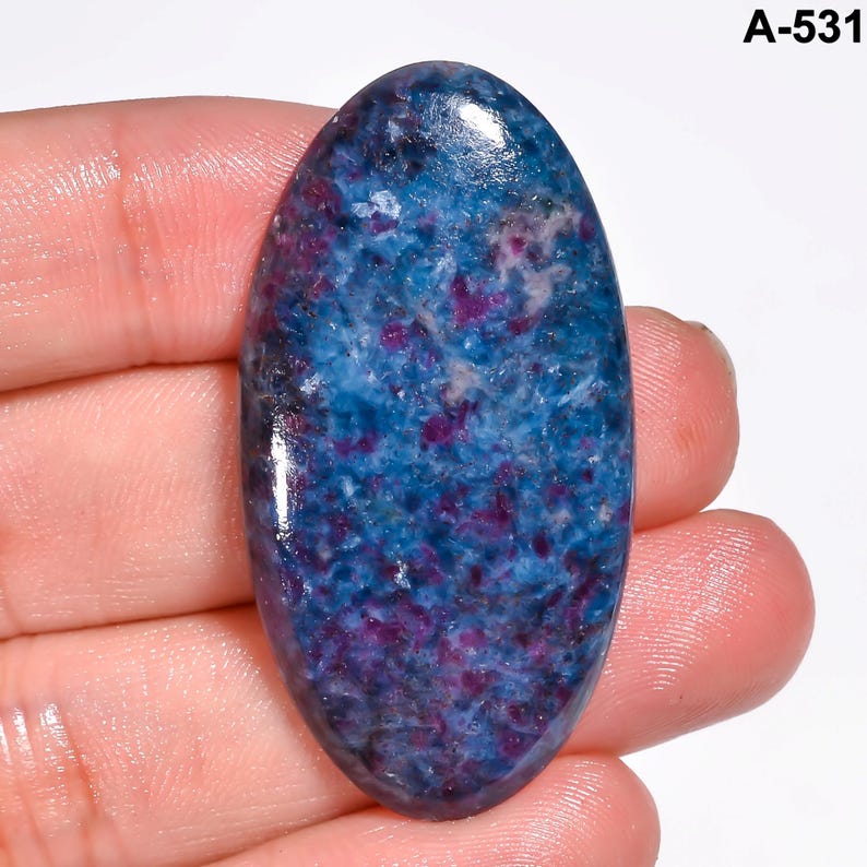 Pode incluir: Uma pedra preciosa ovalada com uma superf&iacute;cie polida. A pedra apresenta uma vibrante mistura de tons azuis e roxos, criando um padr&atilde;o salpicado. A pedra preciosa &eacute; segurada em uma m&atilde;o, mostrando seu tamanho e forma. O texto "A-531" est&aacute; no canto superior direito.