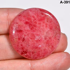 May include: A round, polished gemstone with a vibrant pink hue. The stone has a textured surface with darker red inclusions, and a glossy finish. The gemstone is held in a hand, and the text "A-391" is visible in the upper right corner.