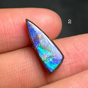 May include: A polished, elongated gemstone with a vibrant display of colors. The stone features a mix of blues, greens, and purples, with a dark border. The number "2" is visible in the background.