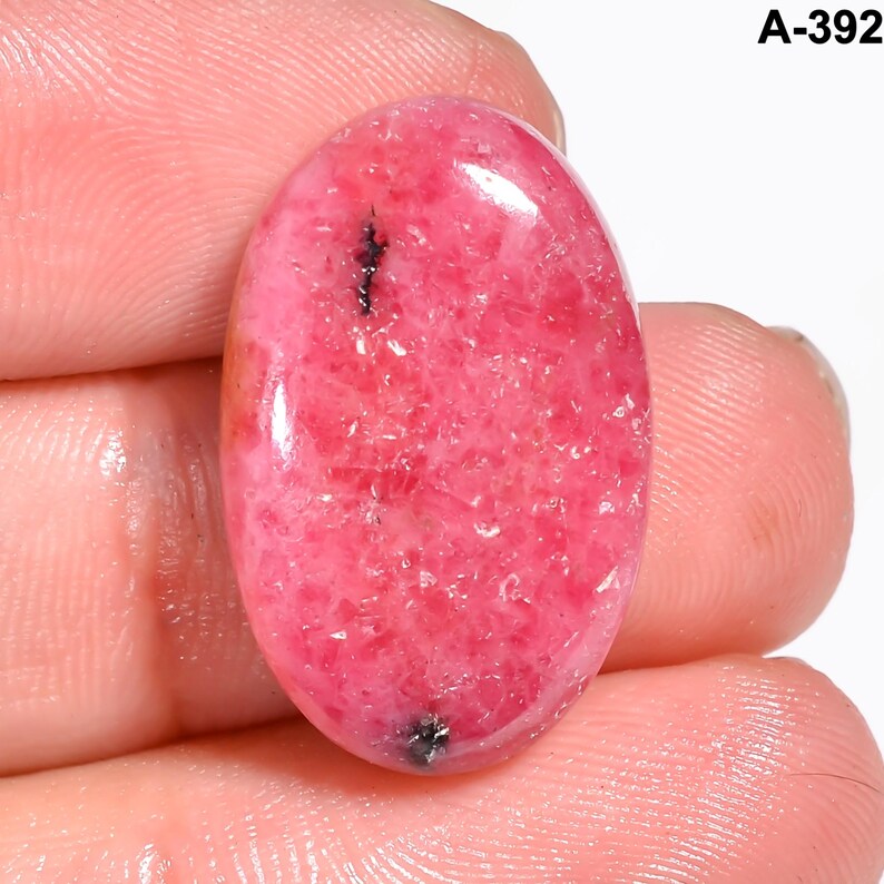 May include: Oval-shaped, polished gemstone in shades of pink with dark inclusions. The stone has a smooth, glossy surface and is held between fingers. The text "A-392" is visible in the upper right corner.