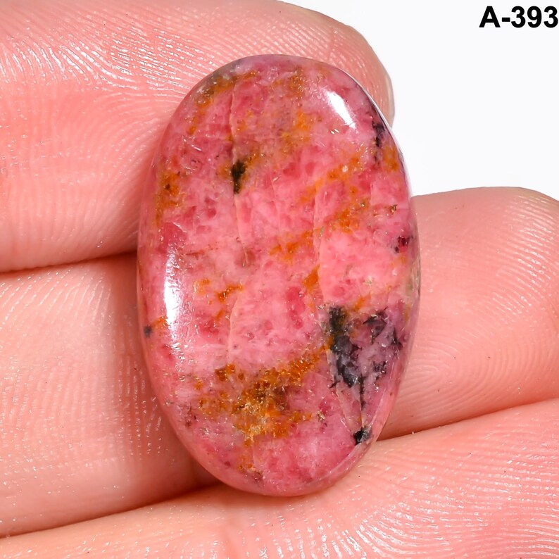 May include: Oval-shaped pink gemstone with orange and black inclusions. The polished stone has a smooth surface and is held in a hand. The text "A-393" is in the upper right corner. This gemstone is suitable for jewelry making.