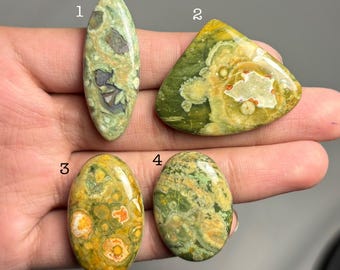 May include: Four polished gemstones in shades of green, yellow, and brown are displayed. The stones vary in shape, including oval and triangular. The gemstones have unique patterns and textures, with visible inclusions and variations in color. The image is well-lit, showcasing the details of each stone.