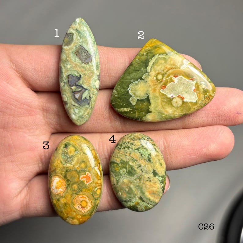 May include: Four polished gemstones in shades of green, yellow, and brown are displayed. The stones vary in shape, including oval and triangular. The gemstones have unique patterns and textures, with visible inclusions and variations in color. The image is well-lit, showcasing the details of each stone.
