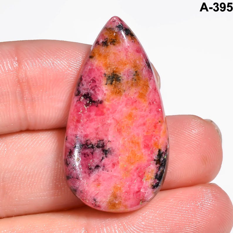 May include: A teardrop-shaped gemstone with a polished surface. The stone features a blend of pink, orange, and black hues, with a marbled appearance. The gemstone is held in a hand, and the text "A-395" is visible in the upper right corner.