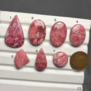 May include: A collection of seven polished gemstones in various shapes, including teardrop, oval, and round. The stones are primarily a deep pink color with white veining. A bronze coin is included for size comparison.
