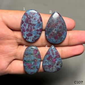 Op de afbeelding: Vier gepolijste cabochon edelstenen met een gevlekt patroon van diep magenta-rode insluitingen in een blauwgrijze matrix. Twee stenen zijn ovaalvormig en twee zijn druppelvormig, elk gelabeld met een wit cijfer van één tot en met vier.