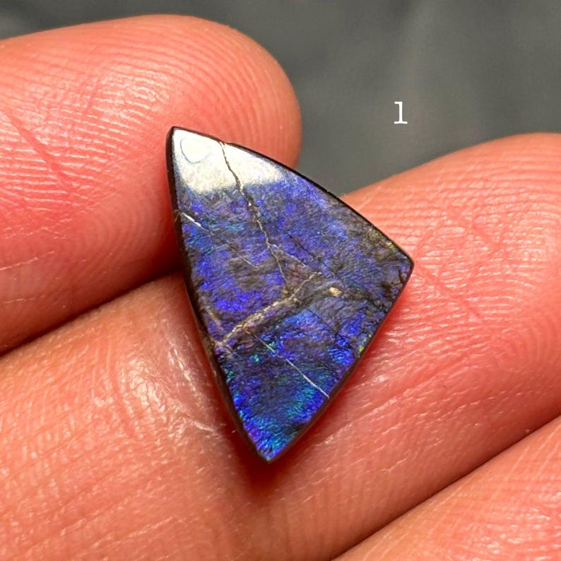 May include: A triangular gemstone with a polished surface. The stone displays a mix of dark blue, purple, and iridescent colors. The number "1" is visible in the background. The gemstone is held in a person's hand.
