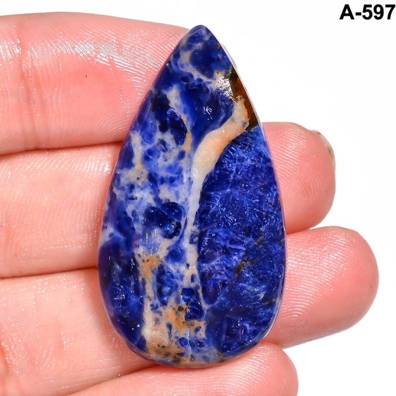 May include: A teardrop-shaped sodalite gemstone with a polished surface. The stone features a striking blend of deep blue, white, and tan hues, with a marbled pattern. The text "A-597" is visible in the upper right corner.