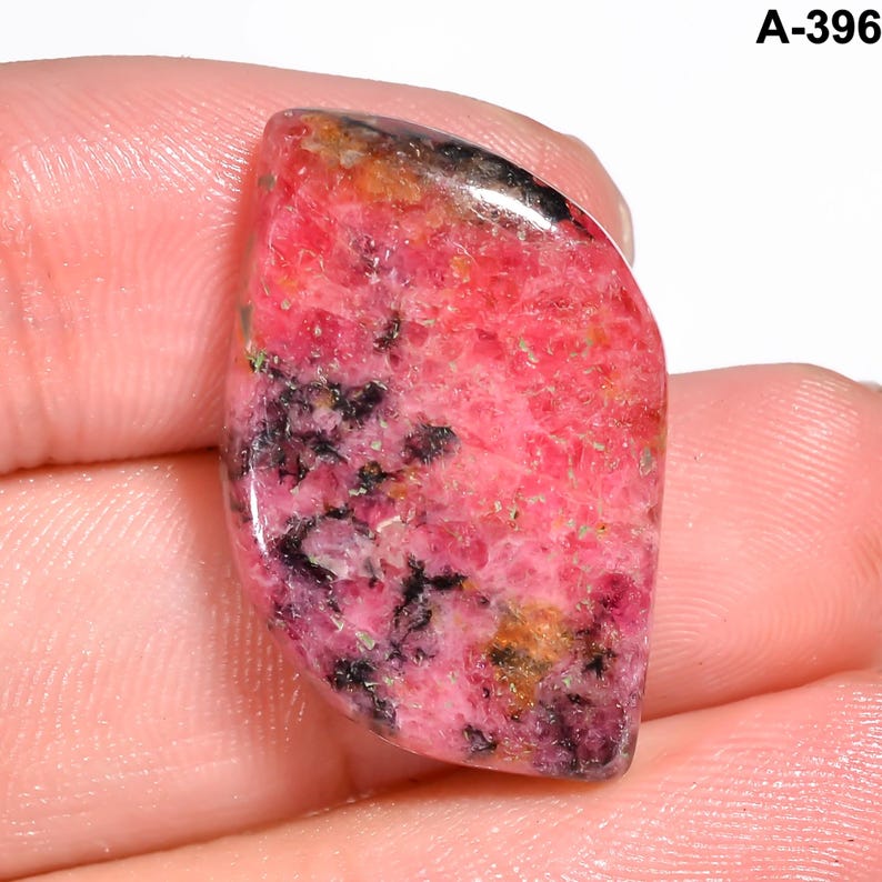 May include: A polished gemstone with a vibrant pink hue, accented by black and gold inclusions. The stone has a smooth, curved shape and is held in a hand. The text "A-396" is visible in the upper right corner.