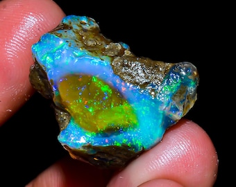 Può includere: Una gemma di opale grezzo dai colori vivaci. La pietra mostra un mix di tonalità blu, verdi e gialle, con lampi di rosso e arancione. L'opale è tenuto in mano su sfondo nero.