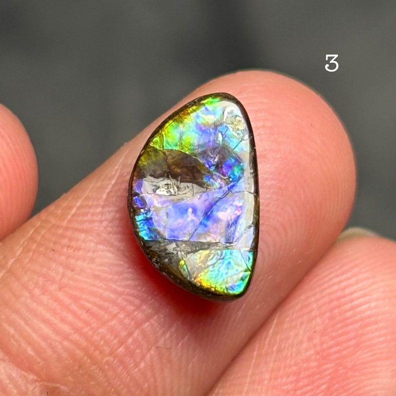 May include: A polished, iridescent gemstone with a vibrant display of colors including blue, green, and yellow. The stone has a dark brown matrix and a smooth, reflective surface. The number "3" is visible in the background.