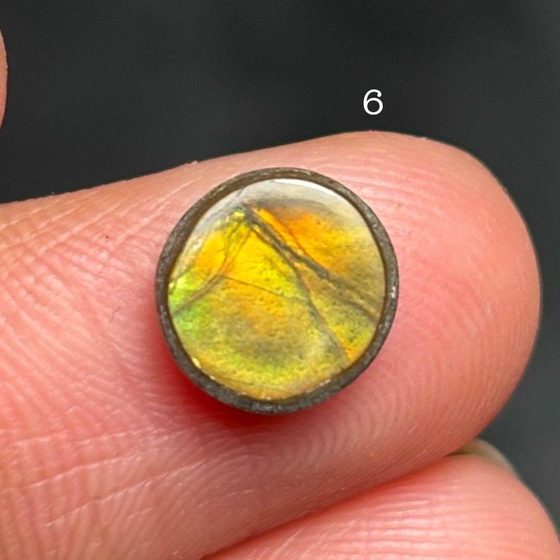 May include: A round gemstone with a vibrant display of yellow, orange, and green hues, encased in a dark metal setting. The gemstone is held between fingers, with the number "6" visible in the background.