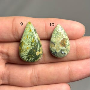 May include: Two teardrop-shaped gemstones with a mix of green, yellow, and brown patterns. The gemstones are held in a hand, with the number "9" and "10" visible above each stone. The stones have a polished surface.