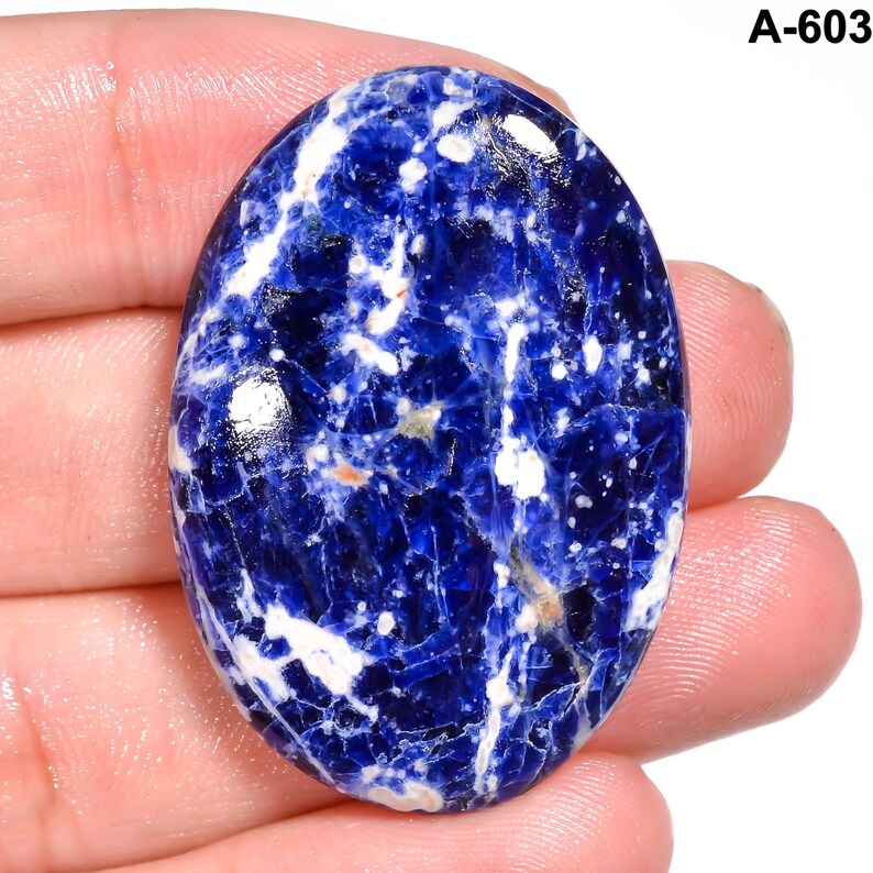 May include: Oval-shaped sodalite gemstone with a deep blue base color and white veining. The stone has a polished surface and is held in a hand. The text "A-603" is in the upper right corner.