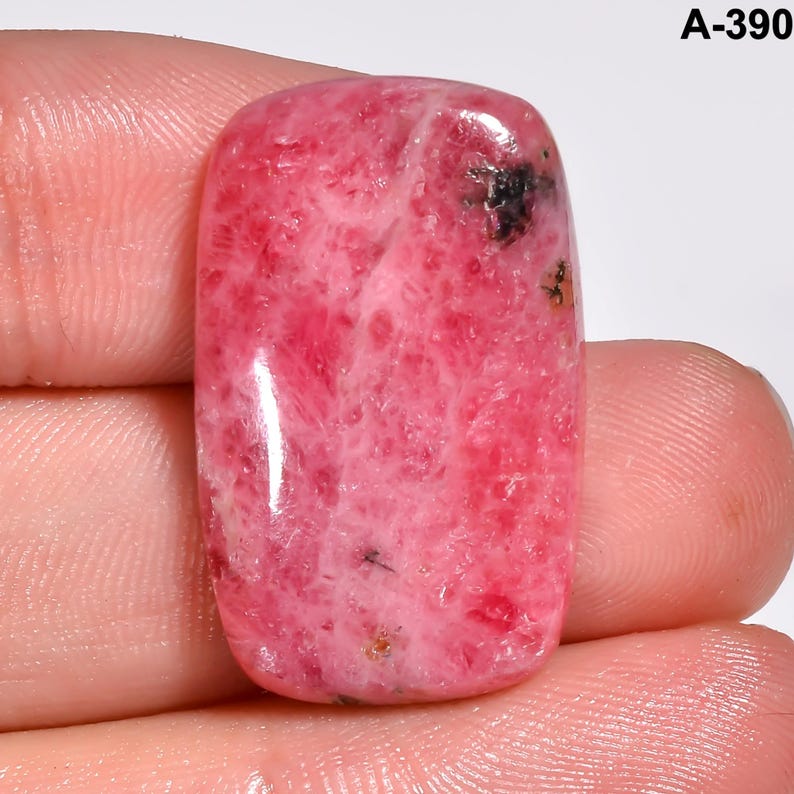 May include: A polished, rectangular gemstone with a vibrant pink hue, featuring a marbled pattern and small dark inclusions. The stone is held in a hand, showcasing its smooth, reflective surface. The text "A-390" is visible in the upper right corner.