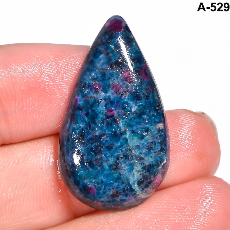 Pode incluir: Uma pedra preciosa em forma de gota, com uma base azul vibrante e inclus&otilde;es vermelhas e roxas. A superf&iacute;cie &eacute; polida e brilhante. O texto "A-529" &eacute; vis&iacute;vel no canto superior direito.