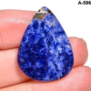 May include: A teardrop-shaped sodalite gemstone with a deep blue hue, veined with white and gray patterns. The stone is held in a hand, showcasing its polished surface and natural variations. The text "A-596" is visible in the upper right corner.