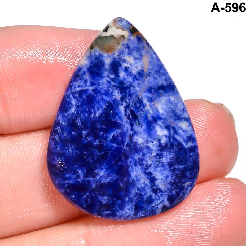 May include: A teardrop-shaped sodalite gemstone with a deep blue hue, veined with white and gray patterns. The stone is held in a hand, showcasing its polished surface and natural variations. The text "A-596" is visible in the upper right corner.