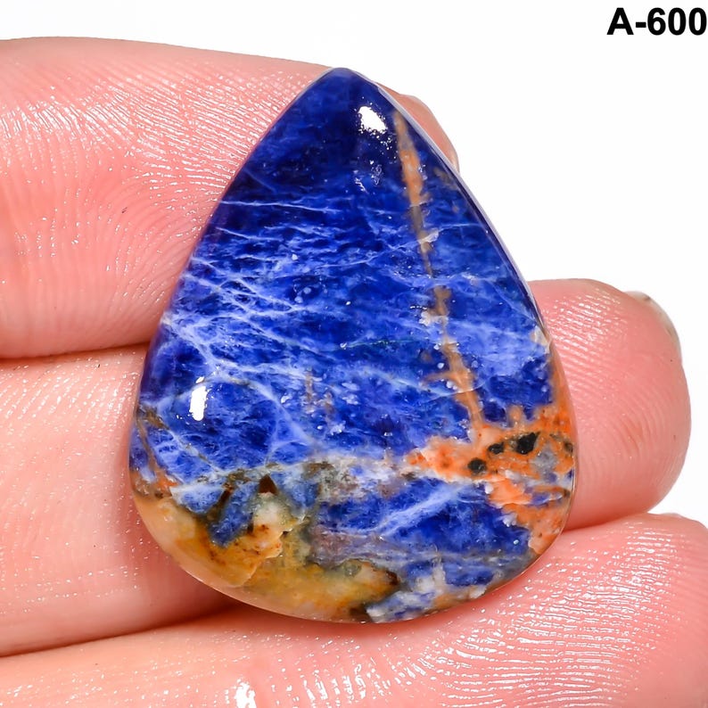 May include: A teardrop-shaped sodalite gemstone with a deep blue hue, veined with white and accented by orange and brown streaks. The stone is polished and held in a hand. The text "A-600" is visible in the upper right corner.