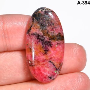 May include: An oval-shaped gemstone with a polished surface. The stone features a blend of colors, including deep pink, black, brown, and orange hues. The gemstone is held between fingers. The text "A-394" is visible in the upper right corner.