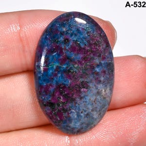Pode incluir: Uma pedra preciosa oval com superf&iacute;cie polida. A pedra apresenta uma mistura de cores, incluindo azul profundo, roxo vibrante e toques de verde. A pedra preciosa est&aacute; rotulada com o texto "A-532" no canto superior direito.