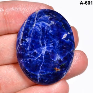 May include: Oval-shaped sodalite gemstone with a deep blue hue, veined with white and light blue streaks. The polished surface reflects light, showcasing the stone's natural patterns. The gemstone is held in a hand.