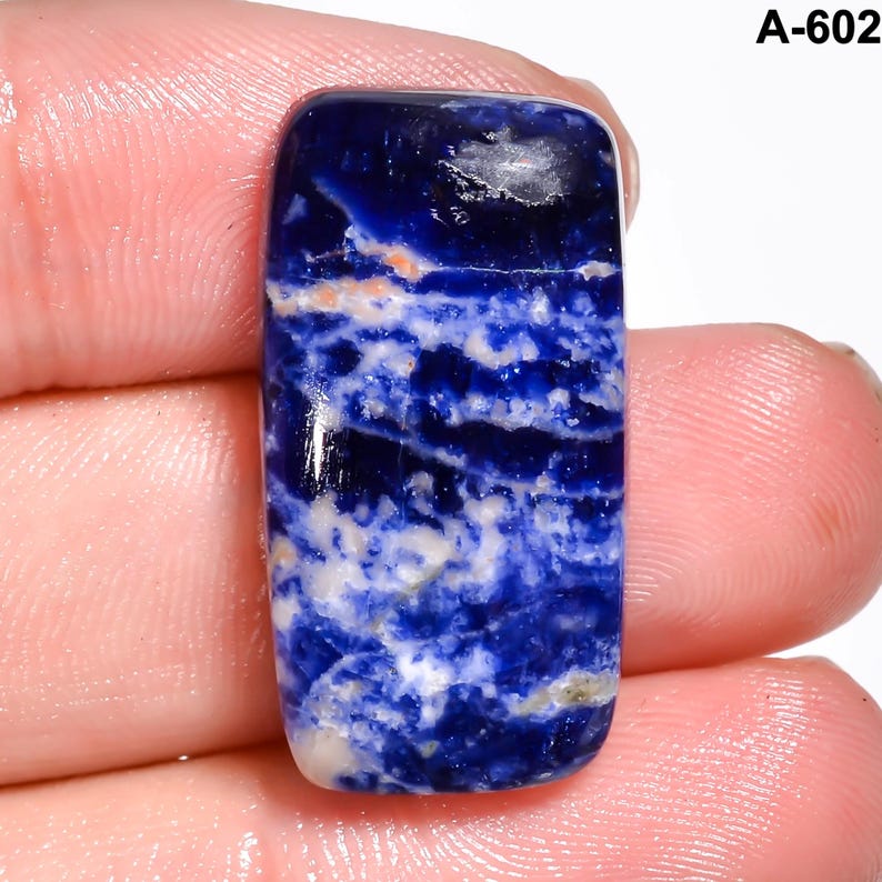 May include: A rectangular, polished gemstone with a deep blue hue, veined with white and hints of orange. The stone is held between fingers, showcasing its smooth, reflective surface. The text "A-602" is visible in the upper right corner.