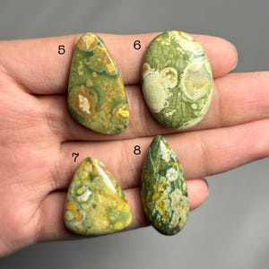 May include: Four polished gemstones in shades of green, yellow, and brown. The stones vary in shape, including oval, teardrop, and triangular. Each stone has unique patterns and textures, with visible inclusions and variations in color.