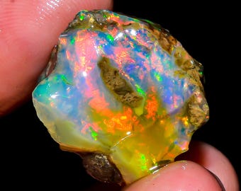 Può includere: Una gemma di opale grezza con una vibrante gamma di colori. La pietra presenta lampi di blu, verde, arancione e giallo. L'opale è tenuto tra le dita, mostrando la sua consistenza naturale, non lucidata e il suo gioco di colori iridescente su uno sfondo scuro.