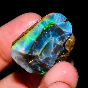 May include: A vibrant, raw opal gemstone with a striking display of color. The stone features a mix of blues, greens, and flashes of orange and red. The opal is held in a hand, showcasing its natural, rough texture and unique patterns.