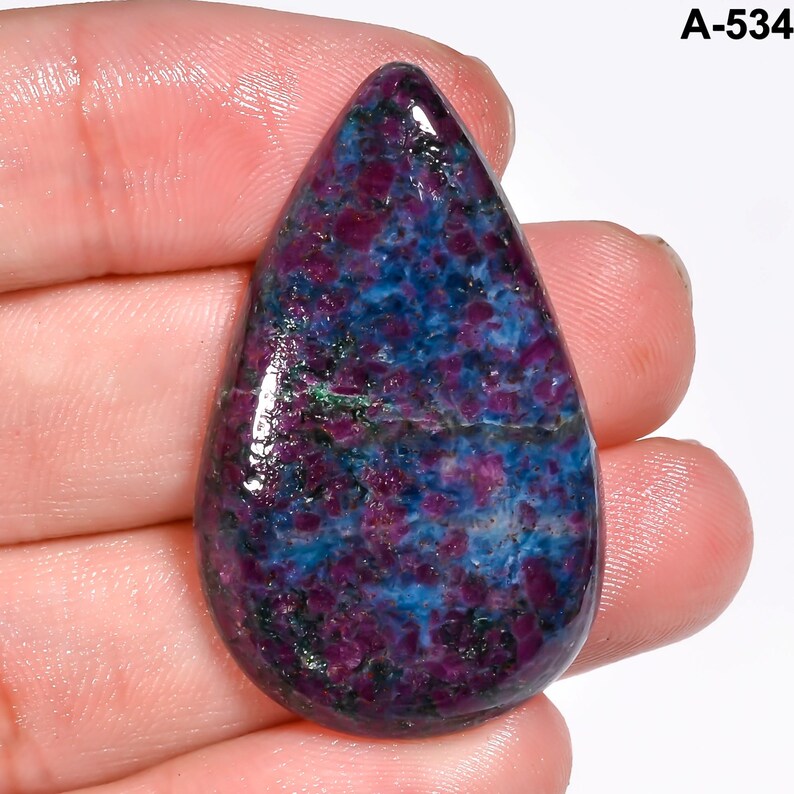 Pode incluir: Uma pedra preciosa em forma de gota com uma superf&iacute;cie polida. A pedra apresenta uma mistura de tons roxos profundos, azuis e verdes, criando um padr&atilde;o salpicado. A pedra preciosa &eacute; segurada em uma m&atilde;o, mostrando seu tamanho e forma. O texto "A-534" est&aacute; no canto superior direito.