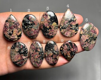 May include: Nine polished cabochons of Eudialyte gemstone, each numbered one through nine, featuring a mottled pattern of deep red, black, and grey mineral inclusions. The stones vary in shape, including teardrop, oval, and diamond-like forms.