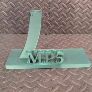 May include: A light teal-colored stand with the letters "MP5" on a rectangular base. An arched support rises from the base. The stand is made of a translucent material and sits on a textured surface.